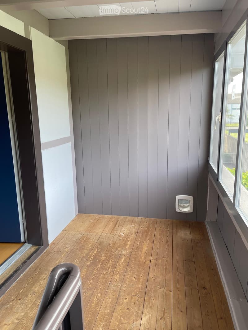 Empty hallway with wooden flooring, grey paneled walls, and large windows on one side, featuring a light switch and a door at the end.