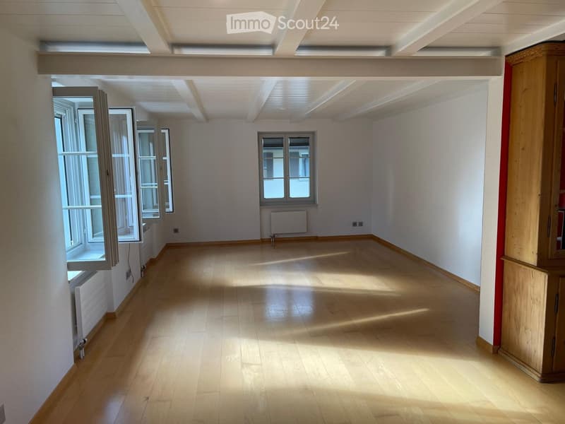 Empty room with wooden flooring, white walls, and large windows allowing natural light to enter. The room features a radiator and electrical outlets, with no furniture or personal items present.