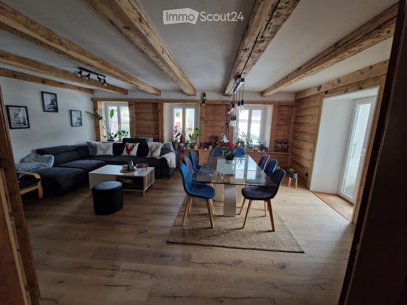 Open-plan living and dining area with wooden beams, exposed wooden floor, large windows, sofa, coffee table, dining table with chairs, and decorative items.