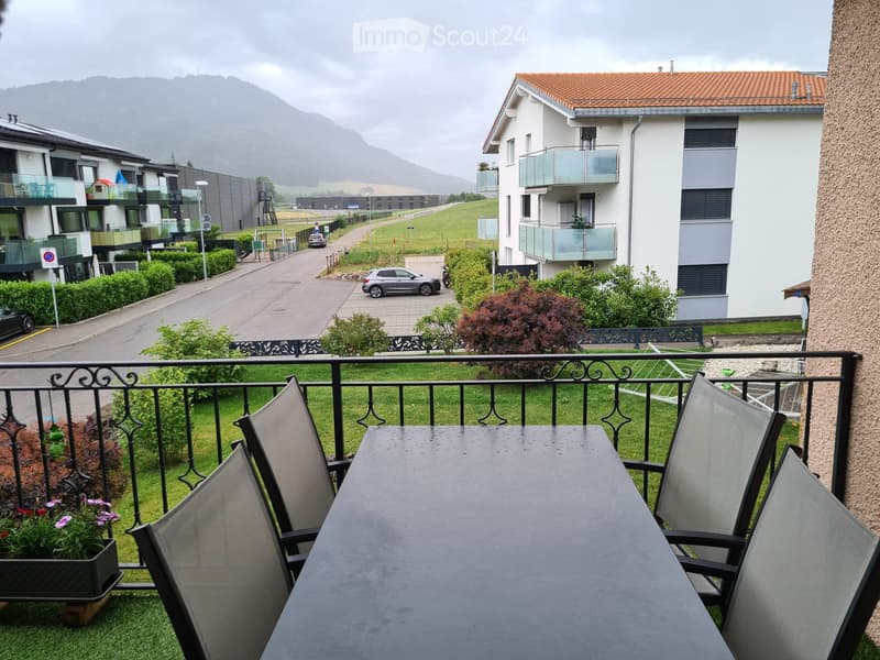 Balcony with a table and chairs, overlooking a landscaped area with buildings and mountains in the background.