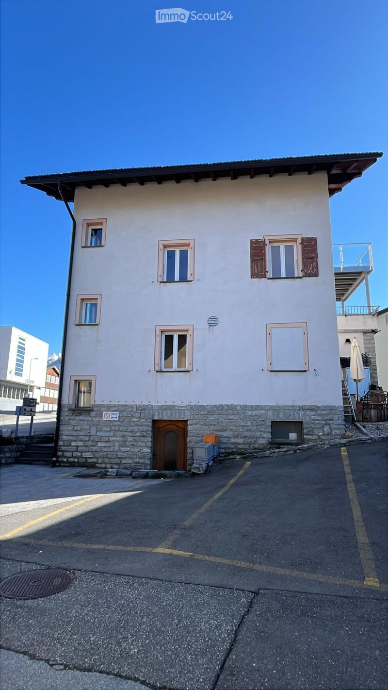 Two-story house with light-colored walls, multiple windows, and a wooden front door. The building has a stone base and a balcony on the upper level.