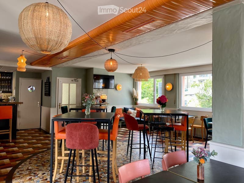 Modern restaurant interior with patterned tile flooring, multiple tables and chairs, pendant lights, and a television mounted on the wall.