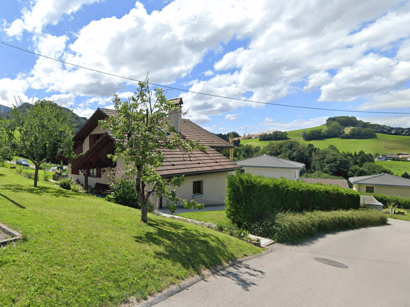 A single-story house with a brown tiled roof, surrounded by greenery and trees, with a scenic mountain view in the background.