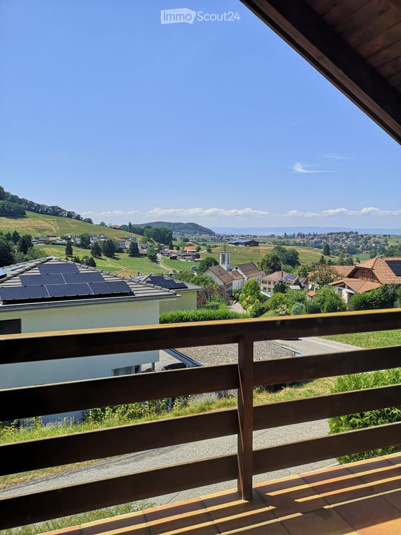 Balcony with wooden railing overlooking a scenic countryside with houses, hills, and solar panels on nearby roofs.