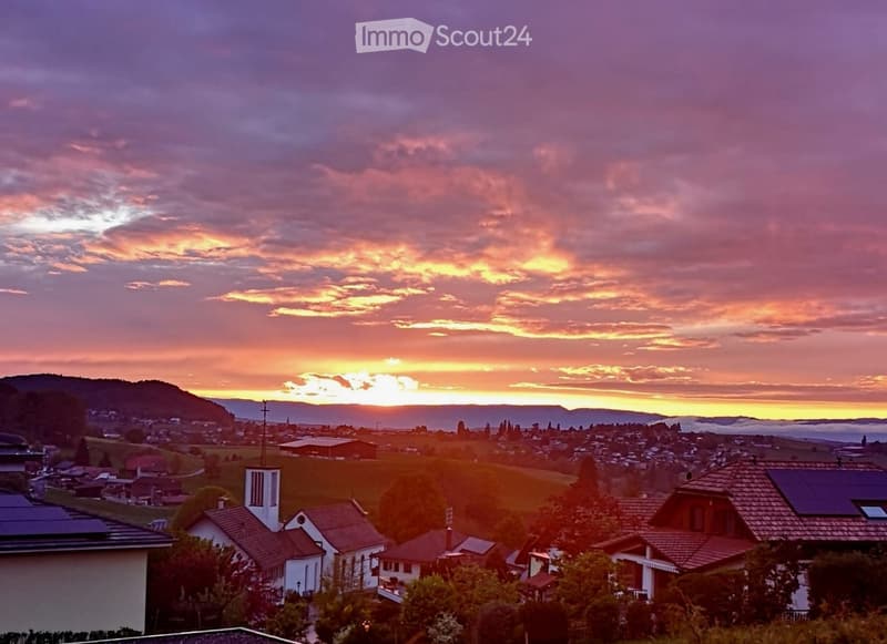 Panoramic view of a town at sunset, with houses, trees, and distant hills under a colorful sky.
