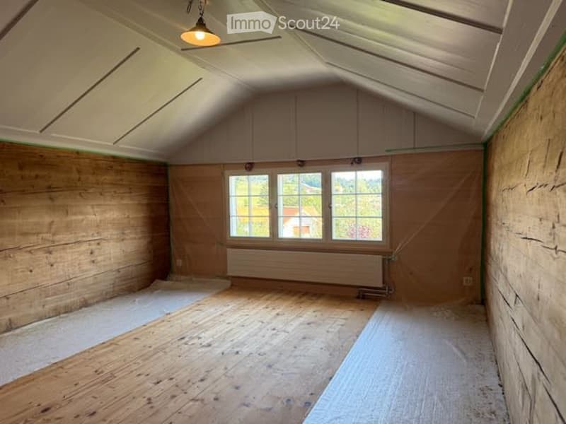 Empty attic room with wooden walls and floor, large window, exposed wooden beams, and a radiator.