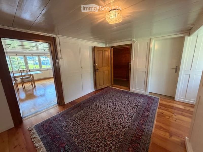 Bright, airy living space with wooden flooring, large area rug, open doorway leading to another room with a dining table and chairs, white walls, and ceiling light fixture.