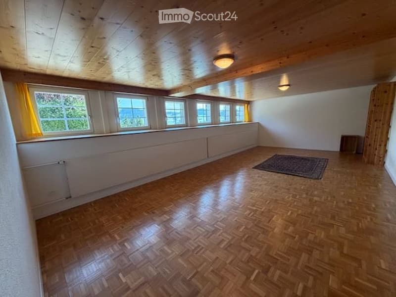 Empty room with wooden floor, white walls, large windows with yellow curtains, exposed wooden ceiling beams, and a small rug in the center.