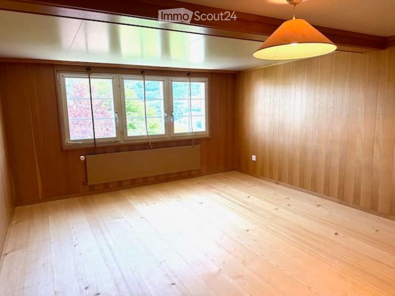 Empty room with wooden walls and floor, large windows with blinds, radiator, and a hanging light fixture.