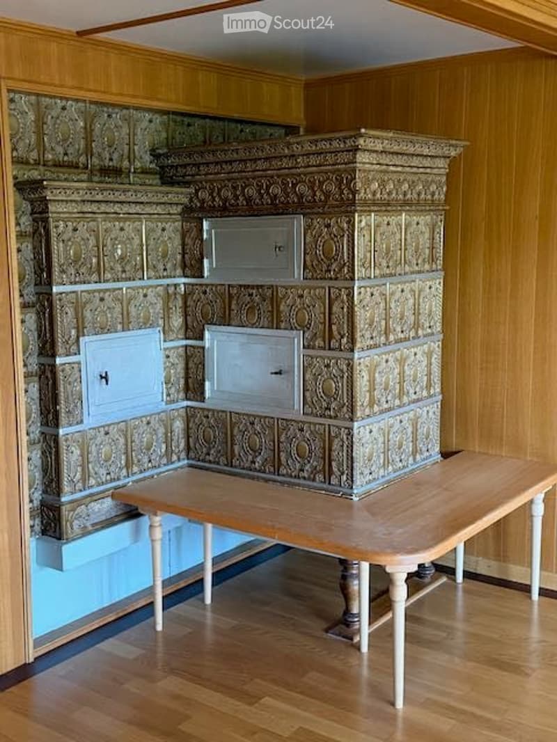 A room with wooden walls and a large, ornate stove. A wooden table and bench are placed in front of the stove.