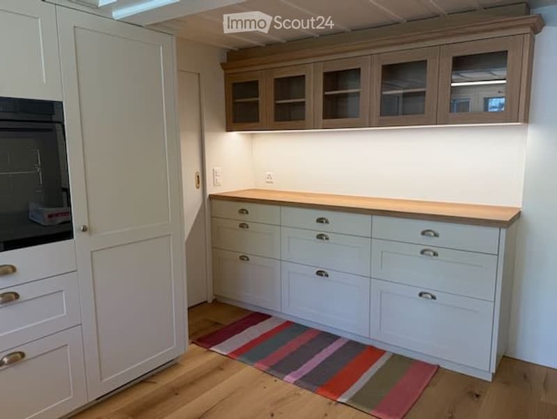 Modern kitchen with light-colored cabinets, drawers, and a countertop, featuring a colorful striped rug on the wooden floor.