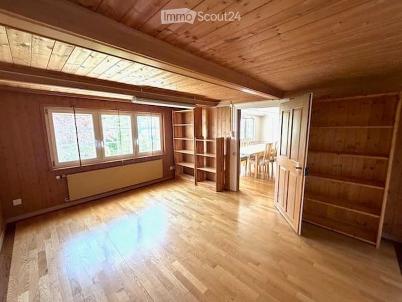 Empty room with wooden walls and floor, large windows, built-in shelving, and an open doorway leading to another room.