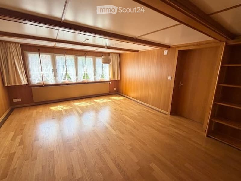 Empty room with wooden flooring, large windows with curtains, wooden walls, and a door leading to another space.