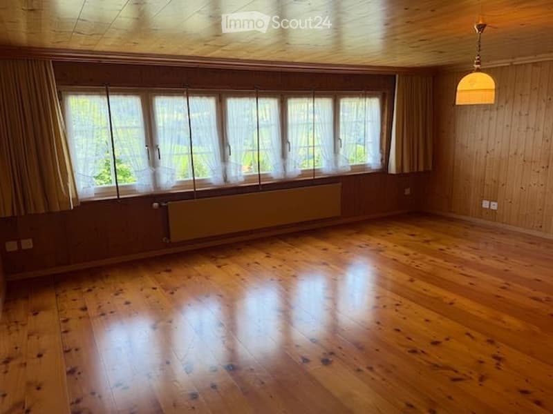 Completely empty room with wooden flooring, large windows with curtains, and a radiator below the windows. No furniture or equipment is present.