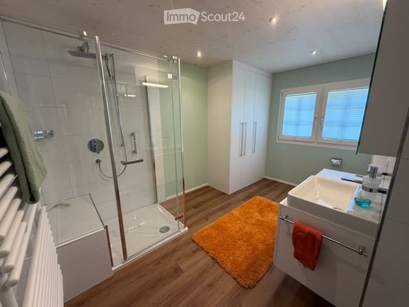 Modern bathroom with a glass-enclosed shower, white tiles, a white sink with a cabinet, and a radiator. An orange bath mat is placed on the wooden floor.