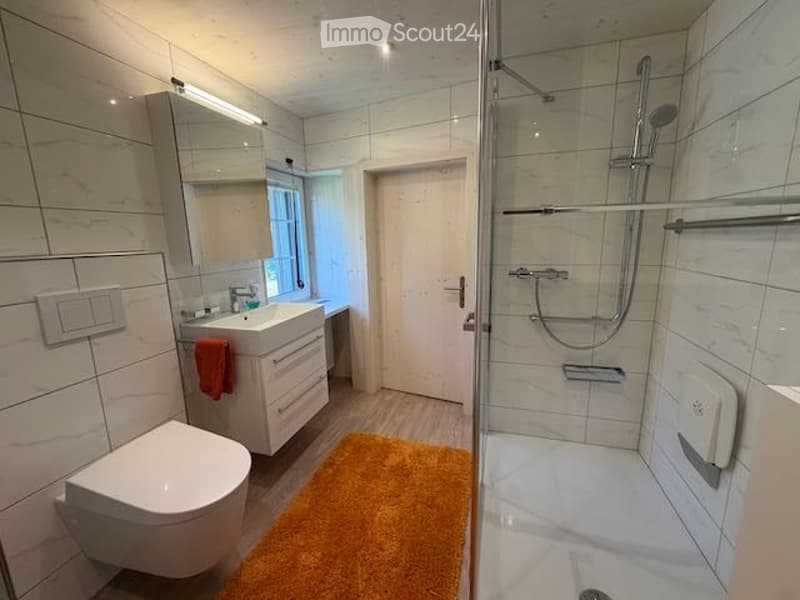 Modern bathroom with white tiles, a toilet, a sink with a mirror, a shower with a glass enclosure, and a door leading to another room.
