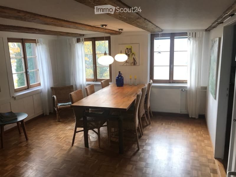 Dining room with wooden floor, large wooden dining table surrounded by chairs, a blue vase, candles, and a small stool with books.