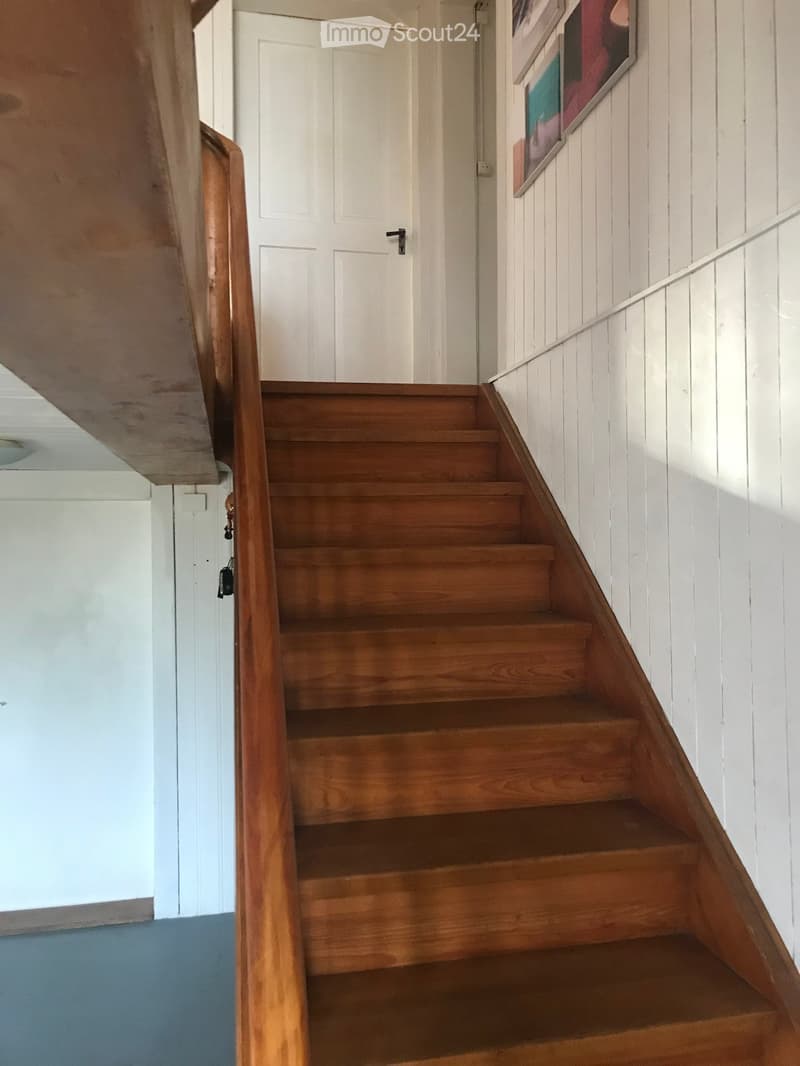 Wooden staircase with white walls and a white door at the top, featuring a wooden handrail and a blue carpeted floor at the bottom.