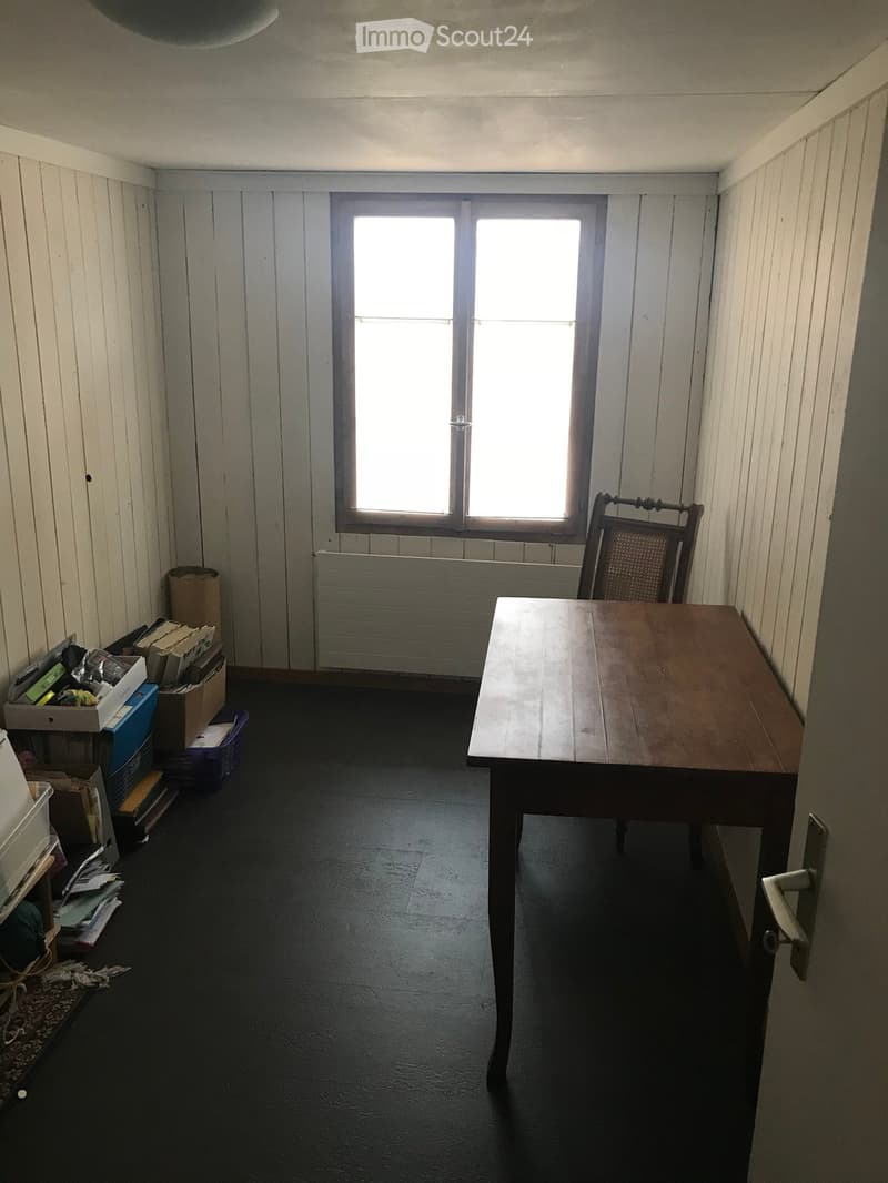 A small, unfurnished room with a wooden table, a single chair, a radiator, and several storage boxes and items stacked against the wall.