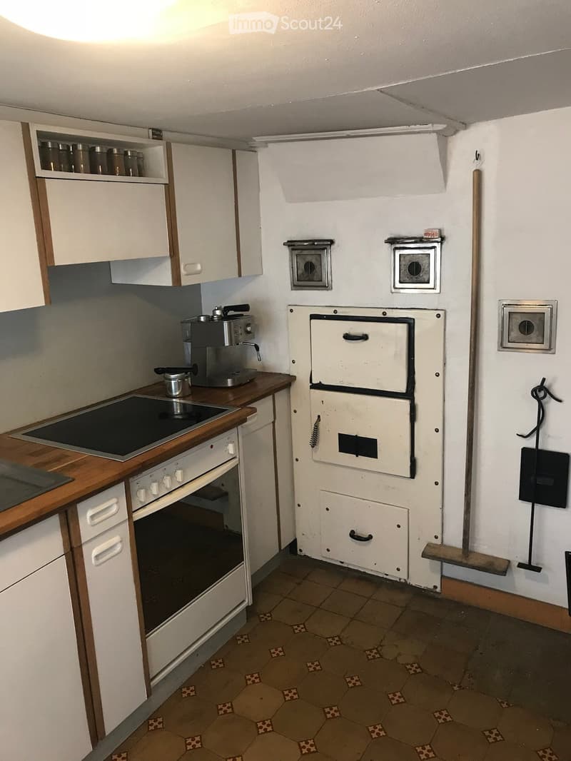 A small kitchen with white cabinets, an induction stove, an oven, and a coffee maker on the counter. The floor is tiled with a hexagonal pattern.