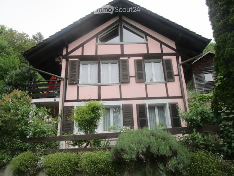 Pink and brown half-timbered house with multiple windows, shutters, and a balcony. Surrounded by greenery and a wooden fence.