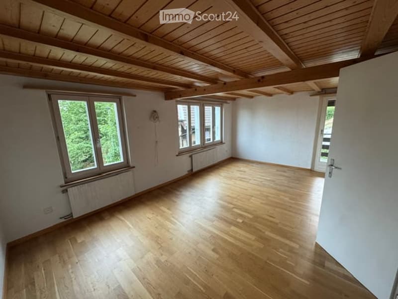 Empty room with wooden flooring, white walls, large windows, and exposed wooden ceiling beams.