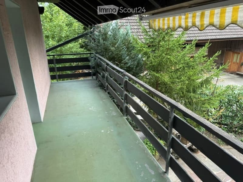 Balcony with green flooring, black metal railing, and a view of surrounding greenery and a neighboring building with a striped awning.