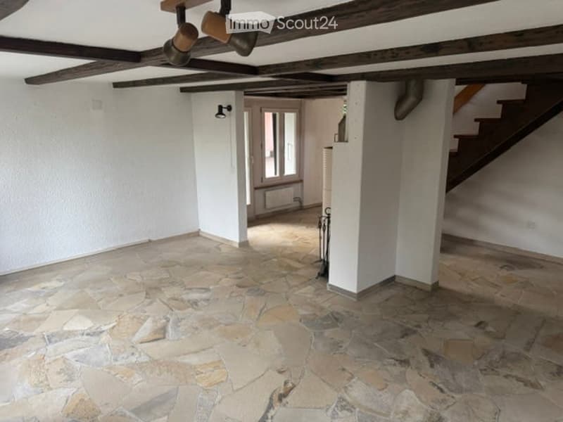 Empty room with exposed wooden beams on the ceiling, stone tiled floor, white walls, and a visible staircase leading upwards.