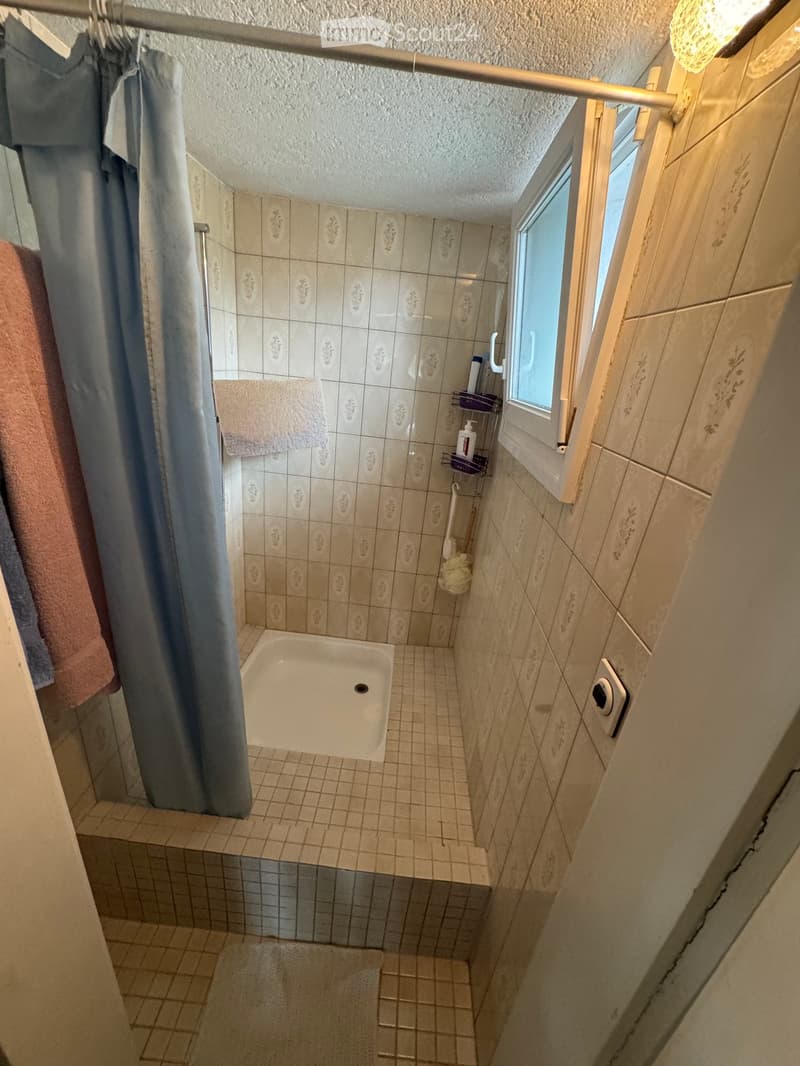 Small bathroom with a bathtub, tiled walls, a window, and a shower curtain. Towels and toiletries are visible on a shelf.