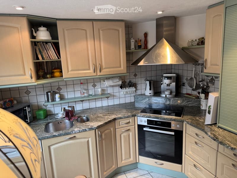 A modern kitchen with light wooden cabinets, granite countertops, and stainless steel appliances including an oven, coffee maker, and microwave. The kitchen features a sink, a kettle, and various kitchen utensils and items on the countertops and shelves.