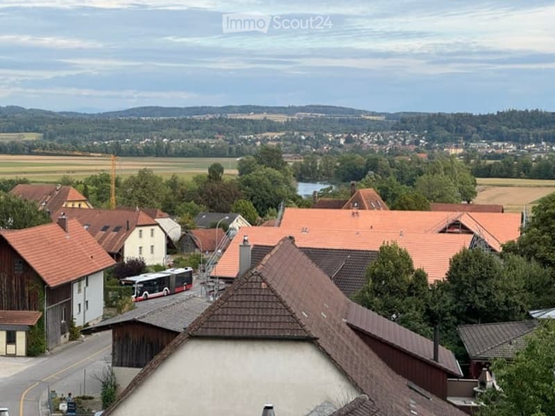 A panoramic view of a small town with red-tiled roofs, scattered houses, and a bus on a road, surrounded by green fields and distant hills.