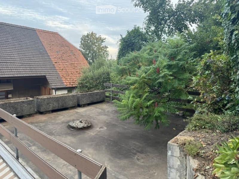 Empty concrete balcony with wooden railing, surrounded by greenery and a small structure with a tiled roof in the background.