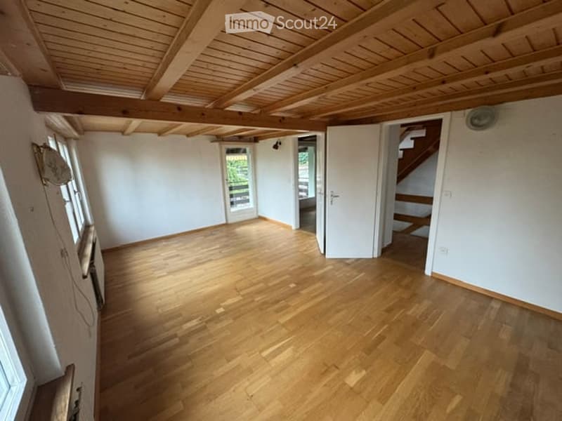 Empty room with wooden flooring and exposed wooden ceiling beams. The room has white walls, large windows, and a door leading to another area.
