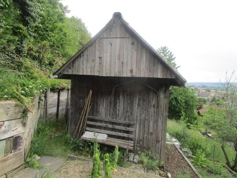 A small wooden shed with a pitched roof, situated in a grassy area with surrounding greenery and a view of a valley in the background.