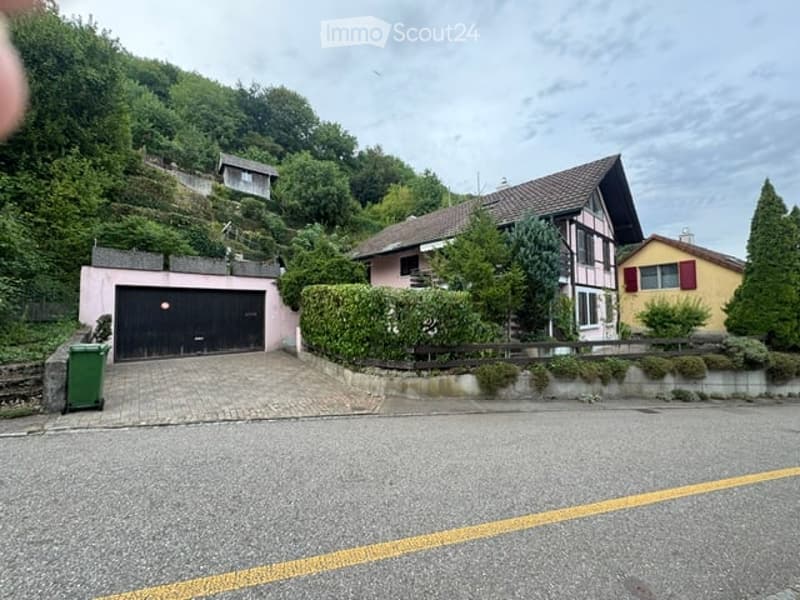 Two-story house with a garage, surrounded by greenery and trees, situated on a hillside.