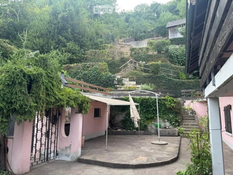 A serene outdoor patio area with a stone floor, surrounded by lush greenery and climbing plants. A small pink structure with a window and a metal gate is visible on the left, while a staircase leads up to a higher level on the right. An umbrella stand and