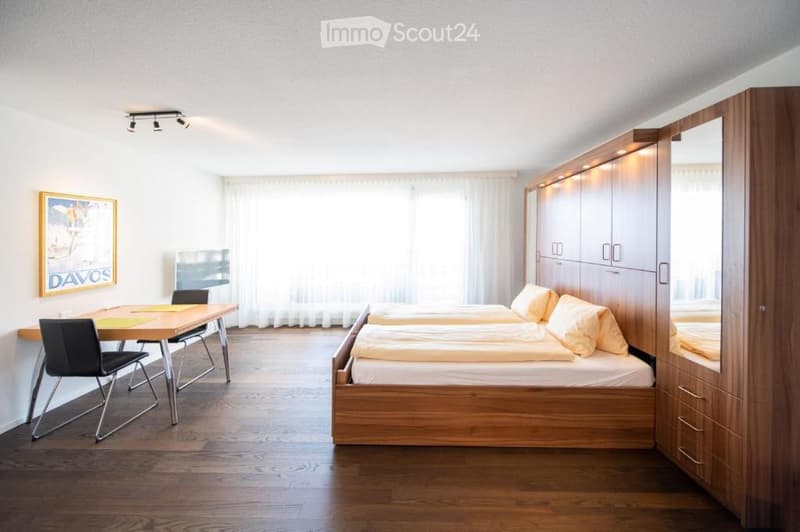 Bright bedroom with wooden flooring, a double bed with pillows and blankets, a wardrobe, a small table with chairs, and a picture frame on the wall.