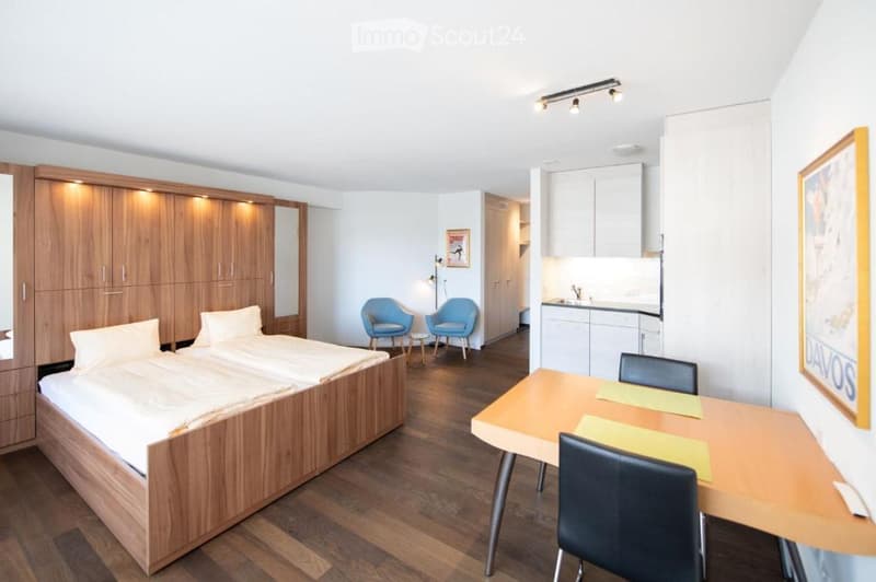 A modern, well-lit bedroom with a large bed, a small sitting area with two blue chairs and a small table, a desk with chairs, and a compact kitchen area with a sink and cabinets.