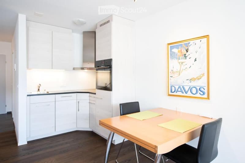 A modern, compact kitchen and dining area with white cabinets, a wooden dining table, black chairs, and a poster on the wall.