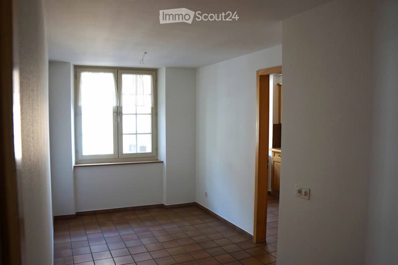 Empty room with tiled floor, white walls, and a window with blinds. A doorway leads to another room with visible furniture.