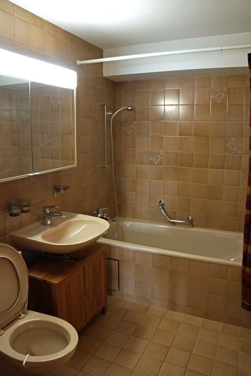 Bathroom with beige tiles, a sink with a faucet, a toilet, a bathtub with a showerhead, and a mirror above the sink.