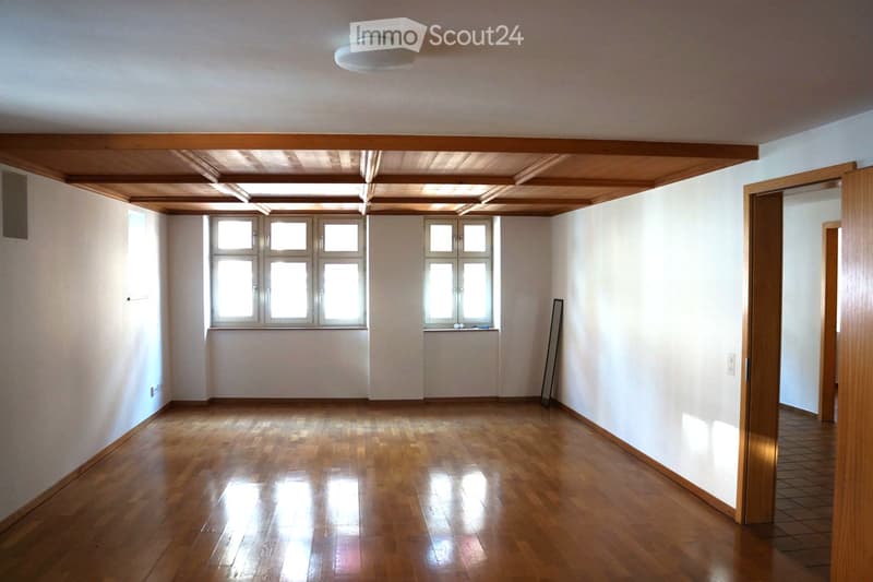 Empty room with wooden flooring, white walls, and large windows allowing natural light. The ceiling features exposed wooden beams, and there is a partially open wooden door leading to another space.