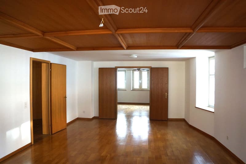 Empty room with wooden flooring, white walls, and wooden doors. Natural light from windows illuminates the space.