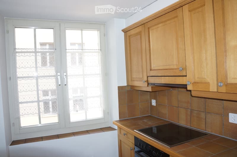 The kitchen features wooden cabinets, a tiled backsplash, and a modern electric stove. There is a large window with white frames and lace curtains, allowing natural light to enter.