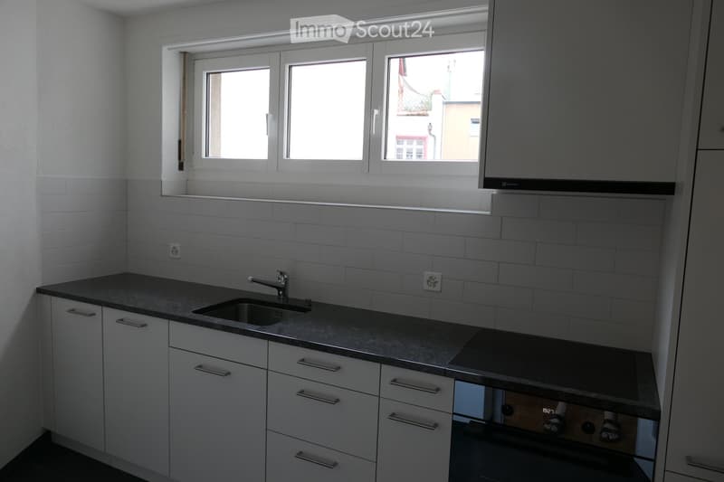 Sleek white cabinets, black countertop, stainless steel sink and faucet, white tile backsplash, oven below countertop, power outlet, window
