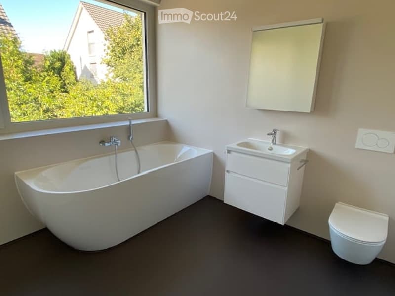 Modern bathroom with a large window offering a view of greenery, featuring a white bathtub, a sink with a cabinet, a toilet, and a wall-mounted mirror.