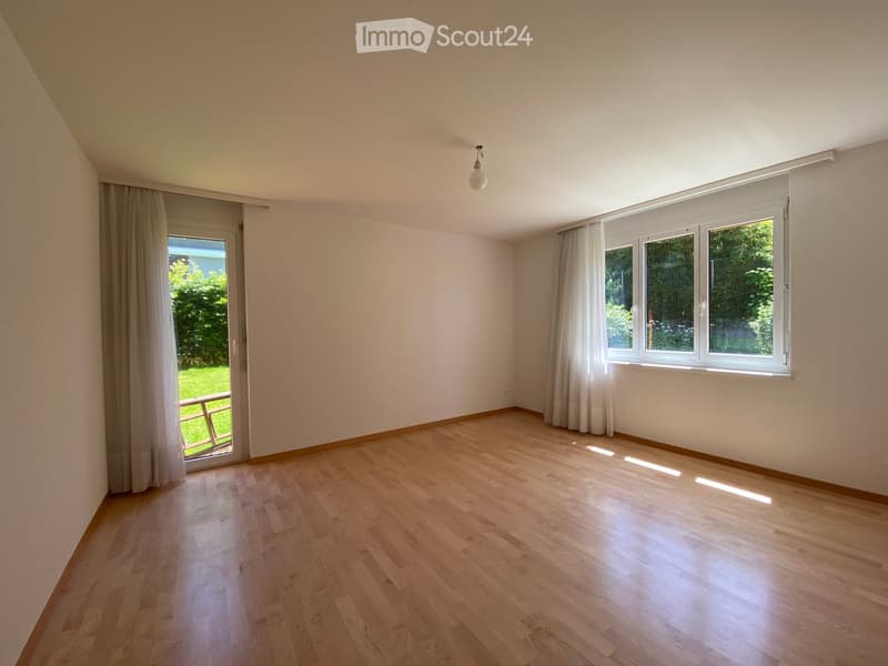 Empty room with wooden flooring, large windows, and a sliding glass door leading to a garden.