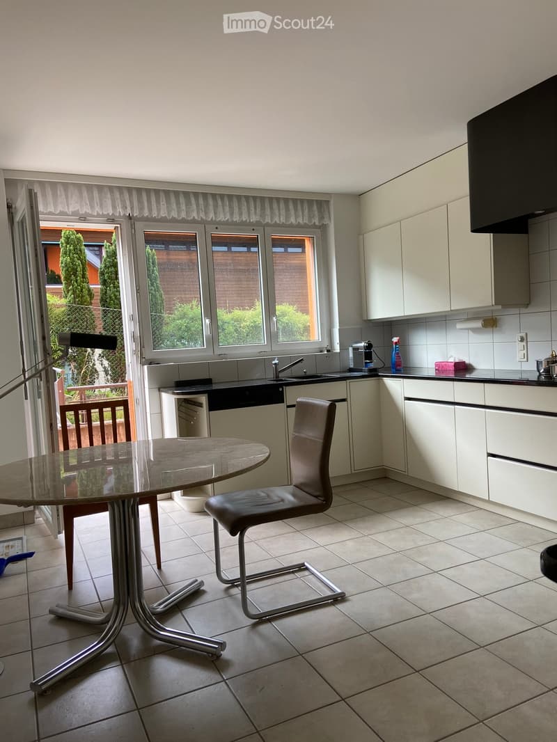 Modern kitchen with white cabinets, tiled floor, round glass table, and a single chair. Features a sink, dishwasher, and various appliances.