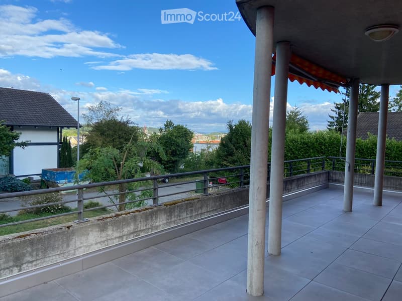 Balcony with tiled floor, metal railing, and columns, overlooking a residential area with trees and houses.
