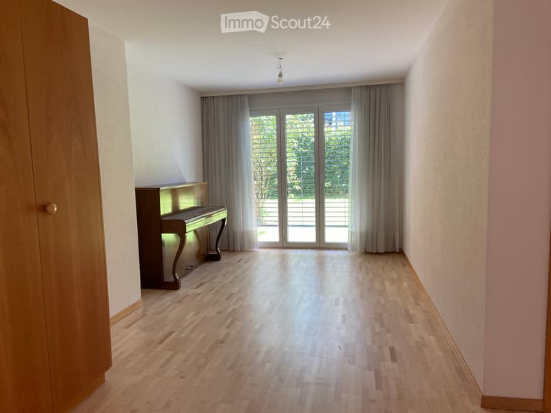 A spacious, well-lit room with wooden flooring, a grand piano, large glass doors with curtains, and a view of greenery outside.